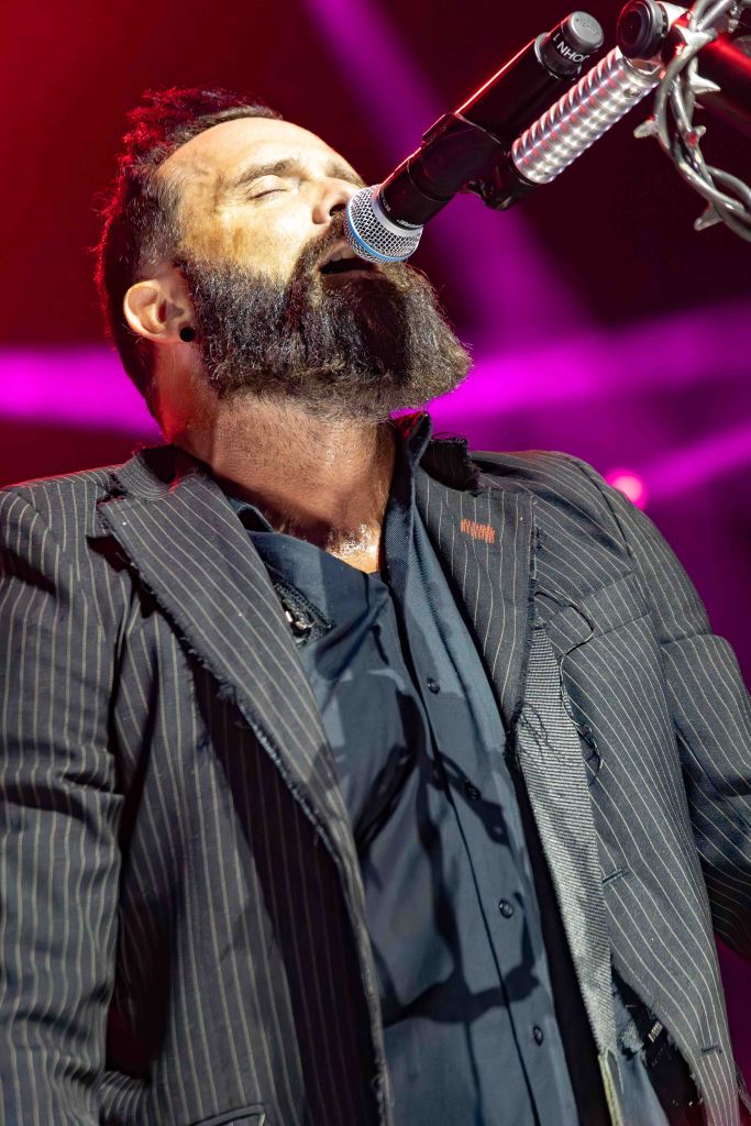 A musician singing passionately into a microphone on stage, wearing a striped suit jacket and a black shirt, with a vibrant light show in the background.