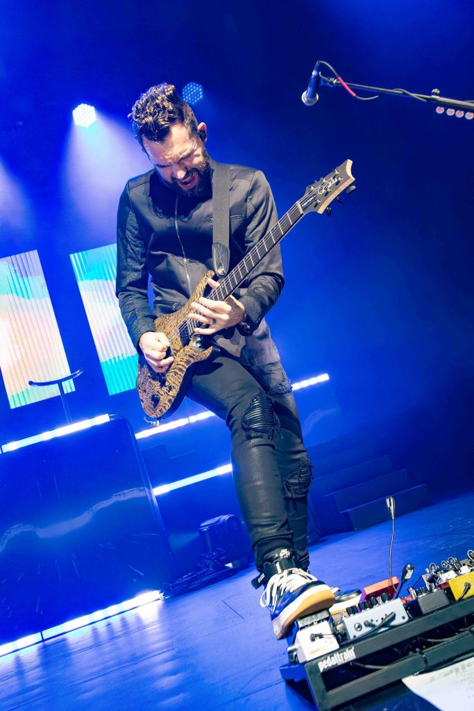 A musician passionately playing an electric guitar on stage, wearing a stylish black outfit and standing on a microphone pedal board, with vibrant blue lighting in the background.