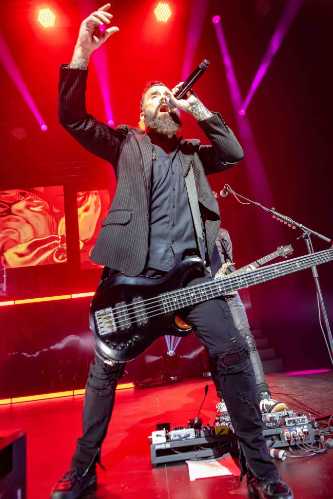 A bearded male singer passionately performing on stage with a black bass guitar, illuminated by dramatic red stage lights.