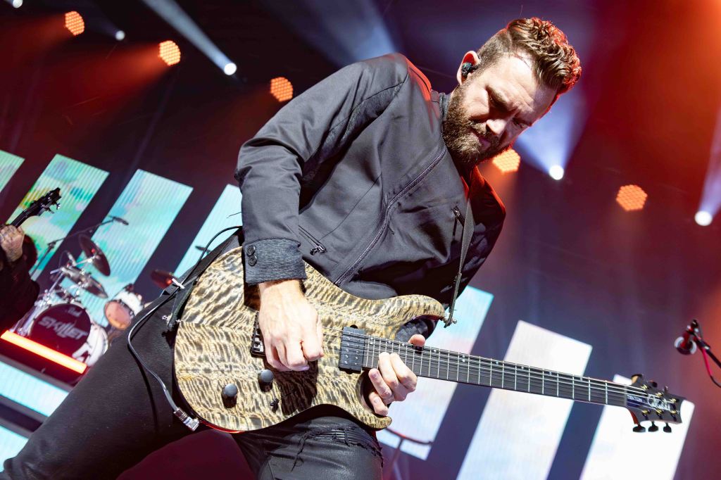 A musician playing an electric guitar on stage during a live performance, with colorful lighting and a band setup in the background.