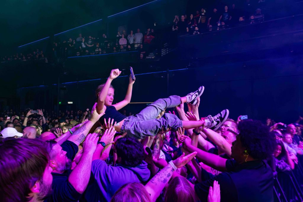 A crowd at a concert supports a person crowd-surfing, hands reaching up to catch and lift them, with vibrant stage lighting in the background.