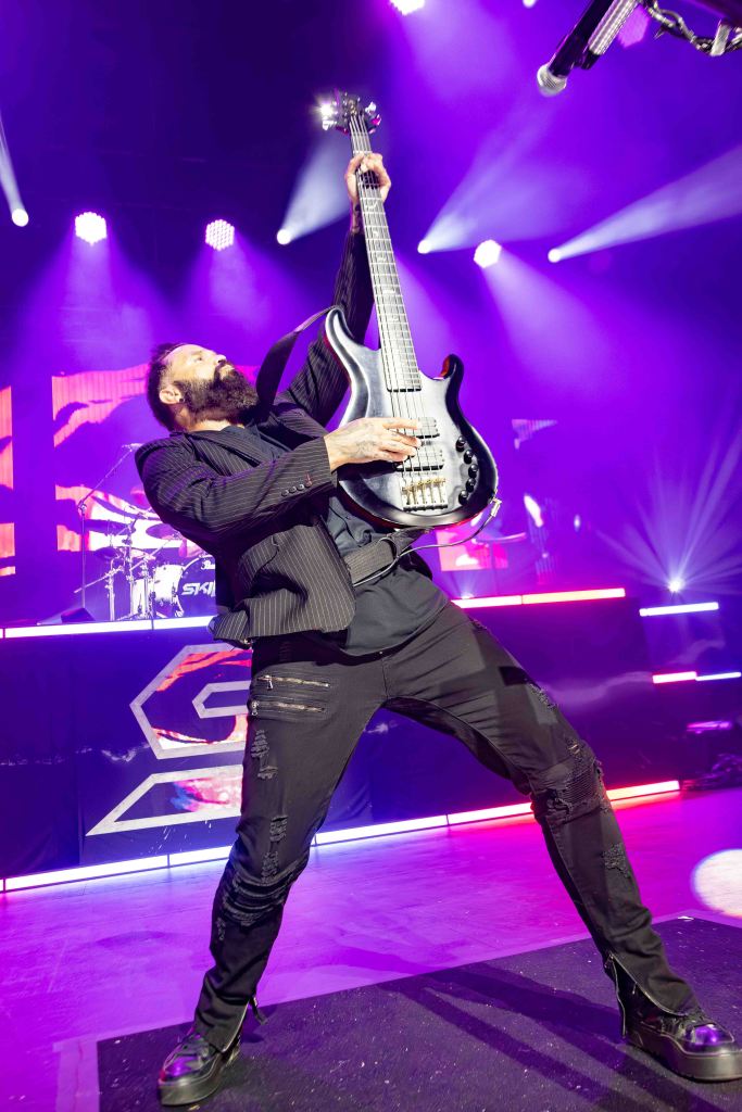 A musician energetically playing an electric bass guitar on stage, dressed in a black suit with ripped pants, against a vibrant backdrop of stage lights.