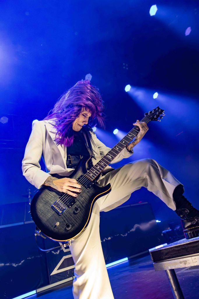 Musician with purple hair performing on stage, playing an electric guitar while wearing a stylish white suit.