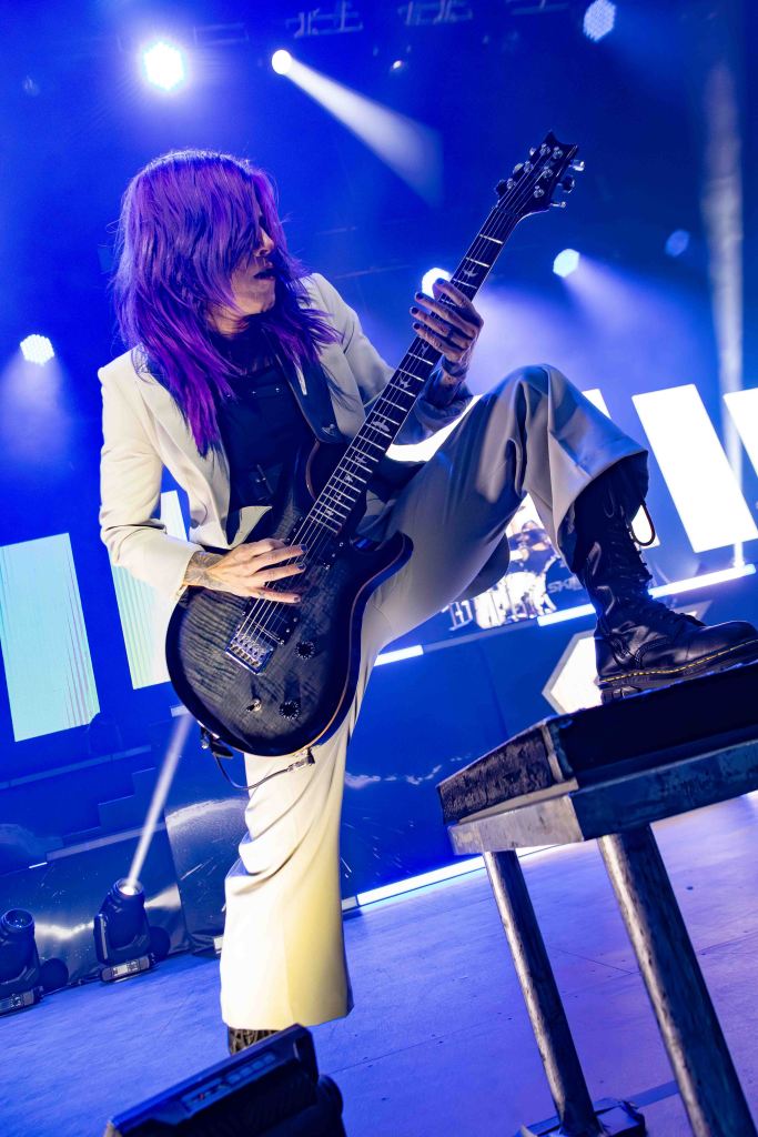 A musician with long purple hair performs on stage, wearing a white suit, and playing a black electric guitar while posing dynamically.