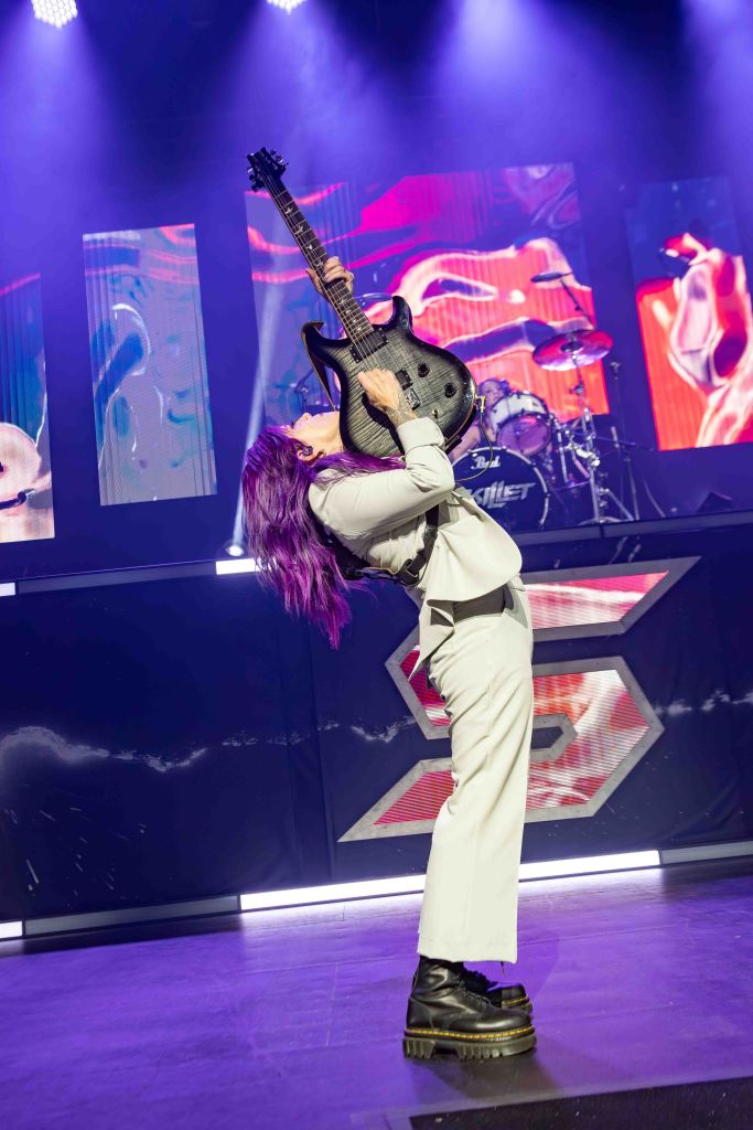 A musician with long purple hair plays an electric guitar on stage, striking a dynamic pose with their head tilted back and the guitar held high, wearing a stylish white outfit and chunky black boots, surrounded by colorful stage lights and a visible drum set in the background.