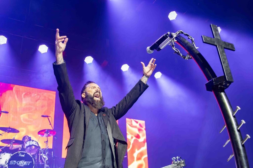 A lead singer performing on stage with arms raised and passionately singing, illuminated by colorful stage lights. In the background, a drummer is visible, and a microphone stand with a cross design is in the foreground.