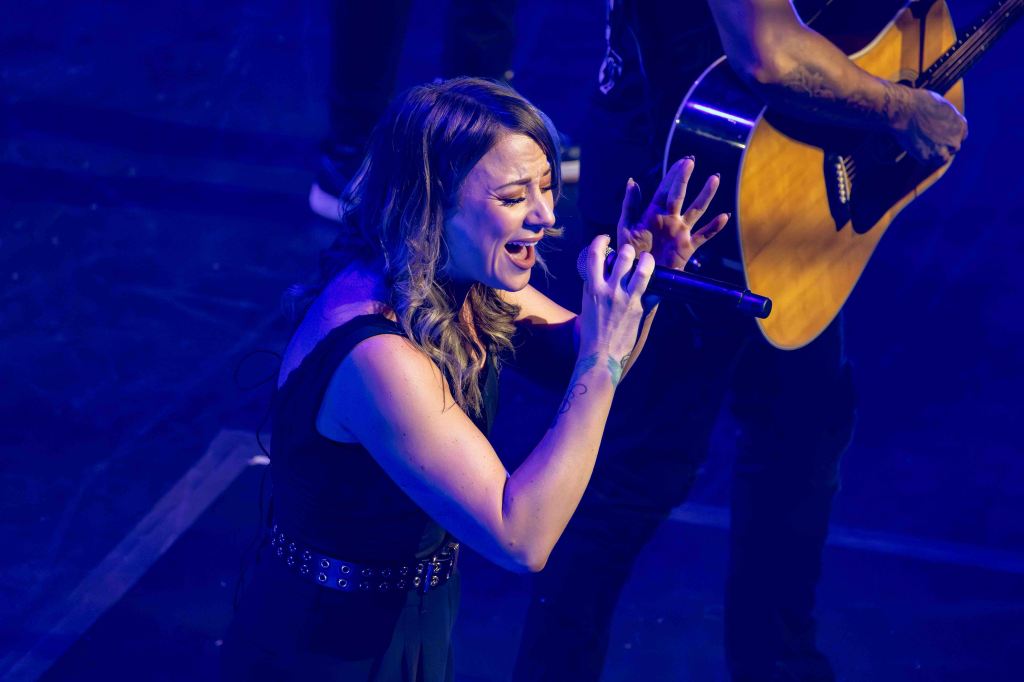 A female singer passionately performing on stage, holding a microphone with a focused expression. She has long, wavy hair and is wearing a black outfit, while a guitarist is visible in the background.