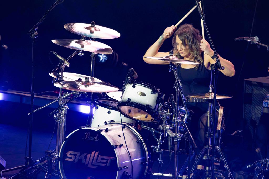A female drummer energetically playing the drums on stage, performing with a band. The drum set features a 'Skillet' logo, and the scene is illuminated with stage lights.
