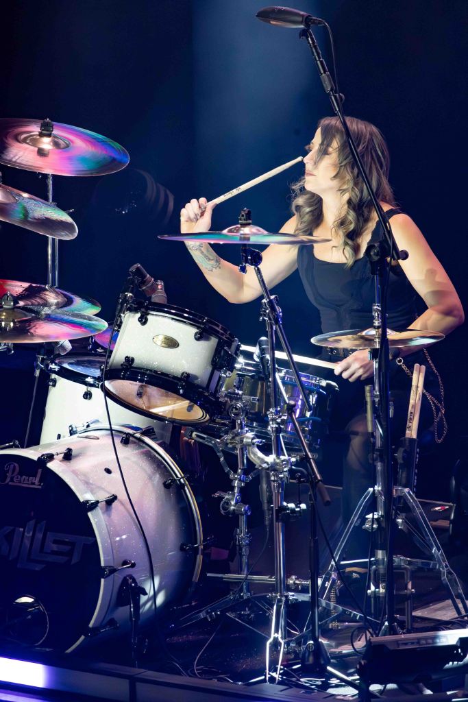 A female drummer performing on stage, energetically playing a drum set with various cymbals, illuminated by stage lights.