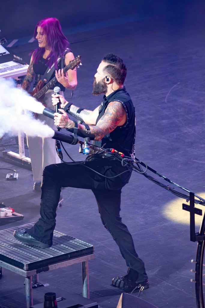 A male musician with a beard performs on stage, holding a microphone and a prop that emits smoke, while a female guitarist plays in the background.
