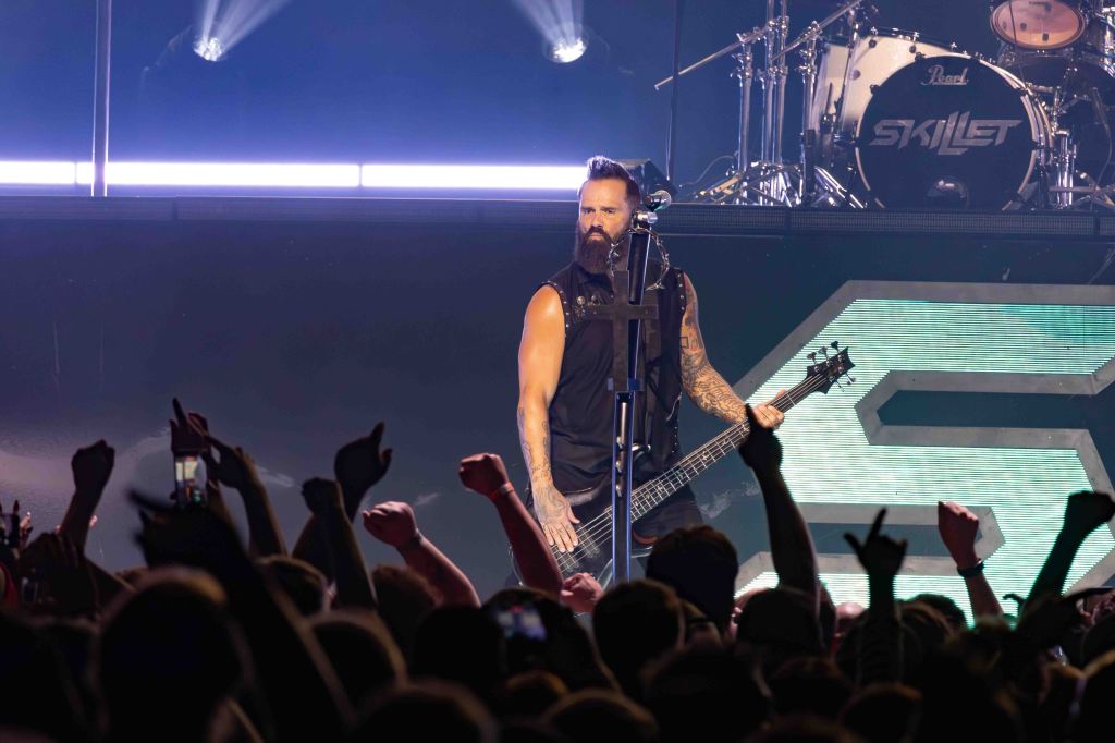 A bassist performing on stage in front of an audience, with dramatic lighting and a drum set in the background.
