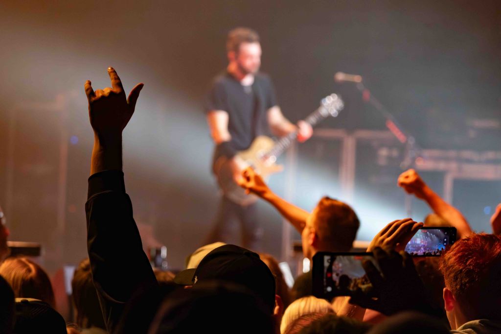 A concert scene featuring a musician playing electric guitar on stage, surrounded by fans with hands raised, some recording with smartphones.