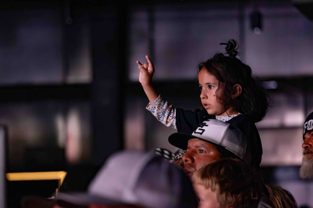 A young girl with dark hair raises her hand enthusiastically while sitting on an adult's shoulders in a crowded setting, possibly at an event or concert.
