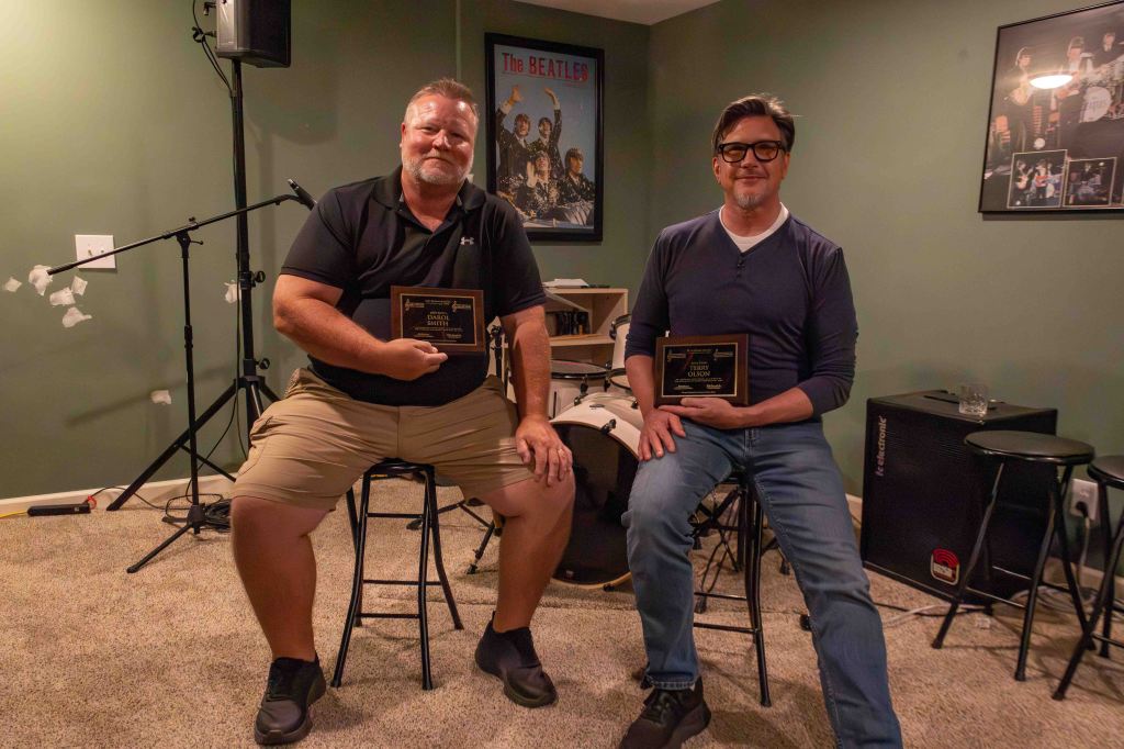 Two men sitting on stools in a music room, each holding an award plaque. The man on the left is wearing a black polo shirt and khaki shorts, while the man on the right is in a dark blue sweater and jeans. Behind them is a drum set and music equipment.