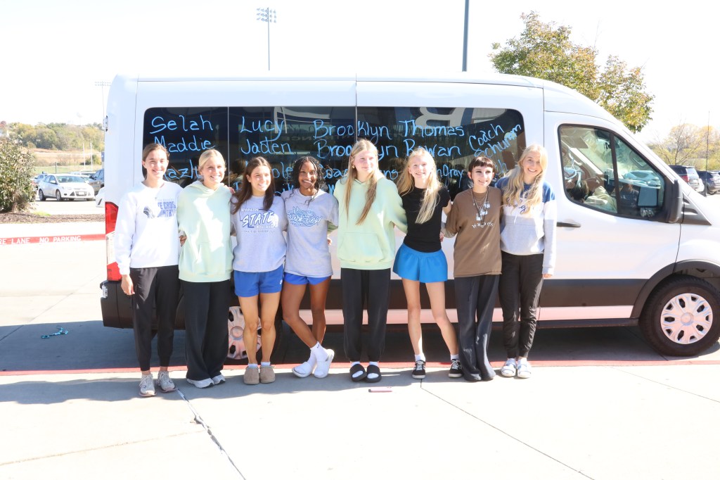 A group of ten female athletes pose together in front of a white van. They are smiling and wearing casual sports attire. The van has names written on its side.