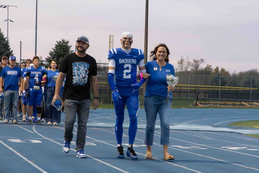 A football player in a blue uniform walks on a track, flanked by two adults, one holding flowers, during a sports event.