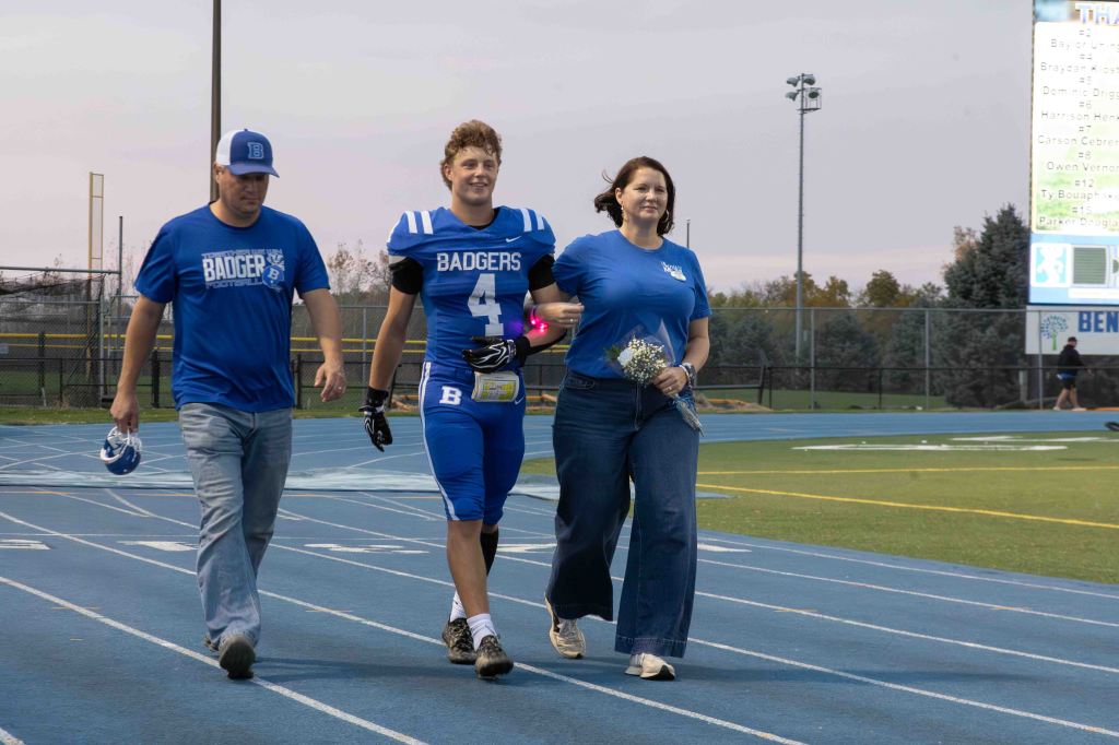 A high school football player in a blue jersey, accompanied by his parents, walks on the track during a senior night celebration. The player carries a bouquet of flowers, while the parents wear blue shirts.