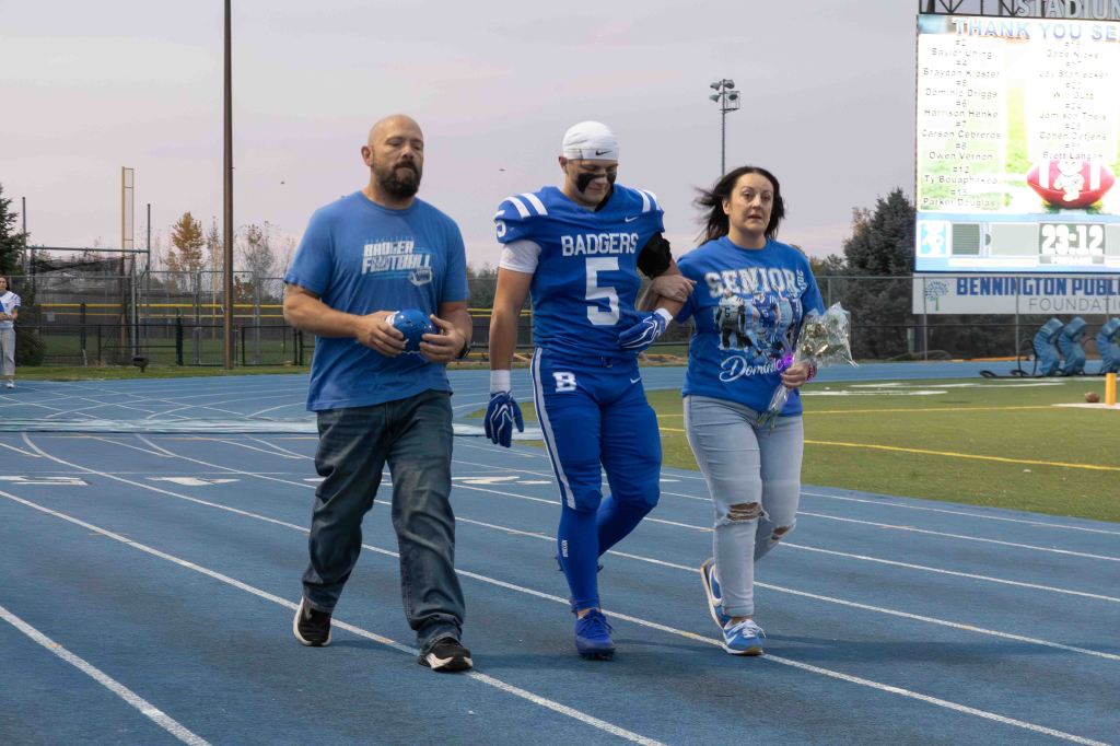 A high school football player in a blue uniform and helmet walks arm in arm with his parents on a football field. The father wears a blue shirt and holds a football, while the mother, dressed in a blue 'Seniors' shirt, carries flowers. The scene takes place during a senior night event under a twilight sky.