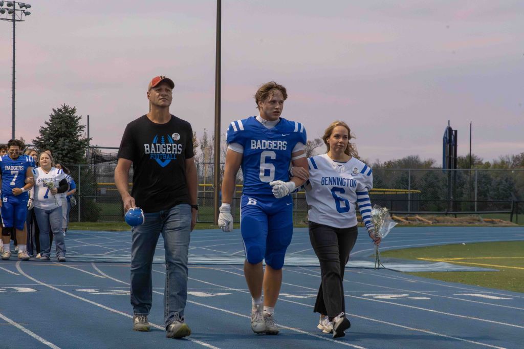 A football player in a blue jersey number 6 walks on a track, accompanied by a man in a black Badgers Football shirt and a woman in a white jersey with blue stripes. They are presenting flowers as part of a ceremony, with other team members visible in the background.