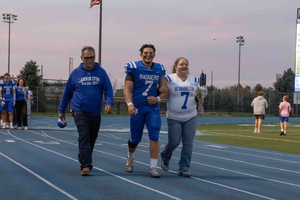 A teenager in a blue football uniform is walking on a track, accompanied by a woman in a white jersey and a man in a blue sweatshirt. They are celebrating a special occasion during a football event at dusk.