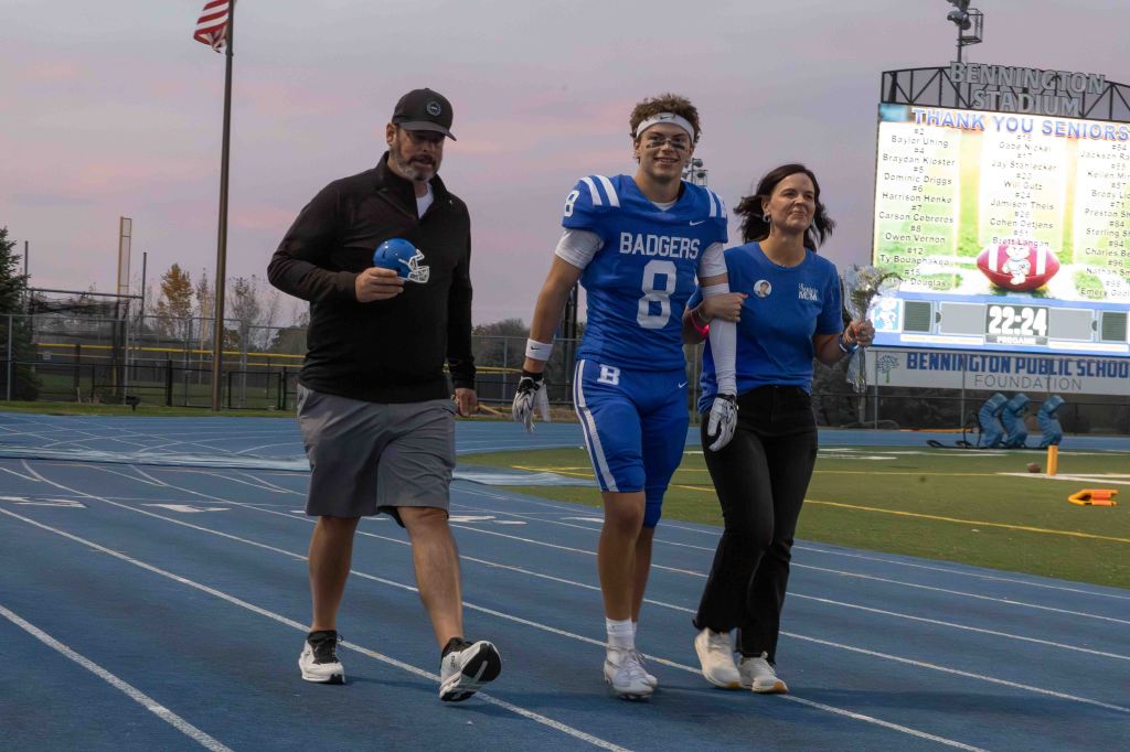 A football player in a blue uniform with the number 8 walks on a track, flanked by a man holding a football helmet and a woman holding flowers, during a senior recognition event at a stadium.