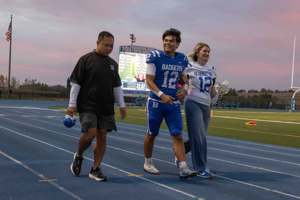A high school football player wearing a blue and white uniform walks on the track with two adults, one in a black shirt and the other in a white shirt, during a sports event at dusk.