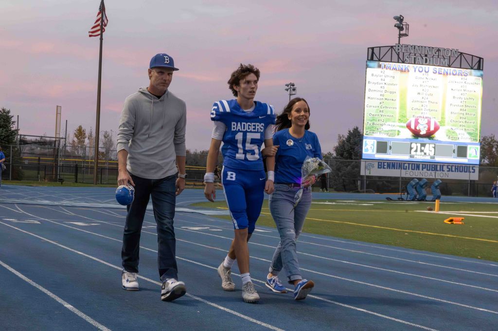 A football player in a blue jersey walks on a track with a woman holding flowers, accompanied by another man, under a twilight sky. A scoreboard displaying 'Thank You Seniors' is in the background.