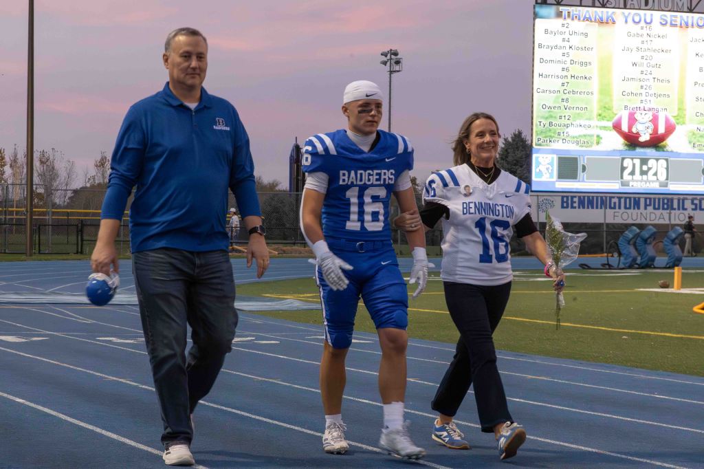 A football player in a blue jersey walks on the track, flanked by a man and a woman. The man wears a blue shirt and holds a football helmet, while the woman wears a jersey and holds flowers. A scoreboard is visible in the background.