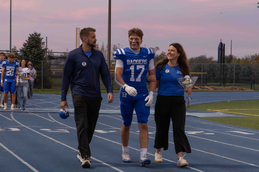 A high school football player in a blue jersey with the number 17 walks on a track, flanked by a man in a blue shirt and a woman in a blue top holding flowers. Other football players and spectators are visible in the background during a celebration event.