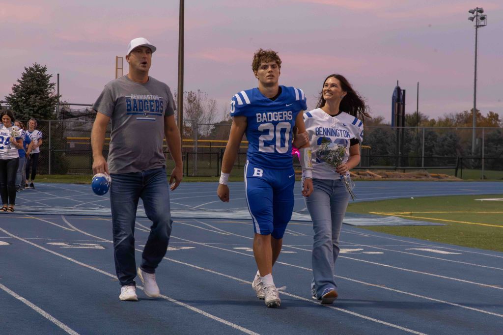 A football player in a blue jersey walks on a track with a woman holding flowers, accompanied by a man in a grey shirt, during a sports event at sunset.
