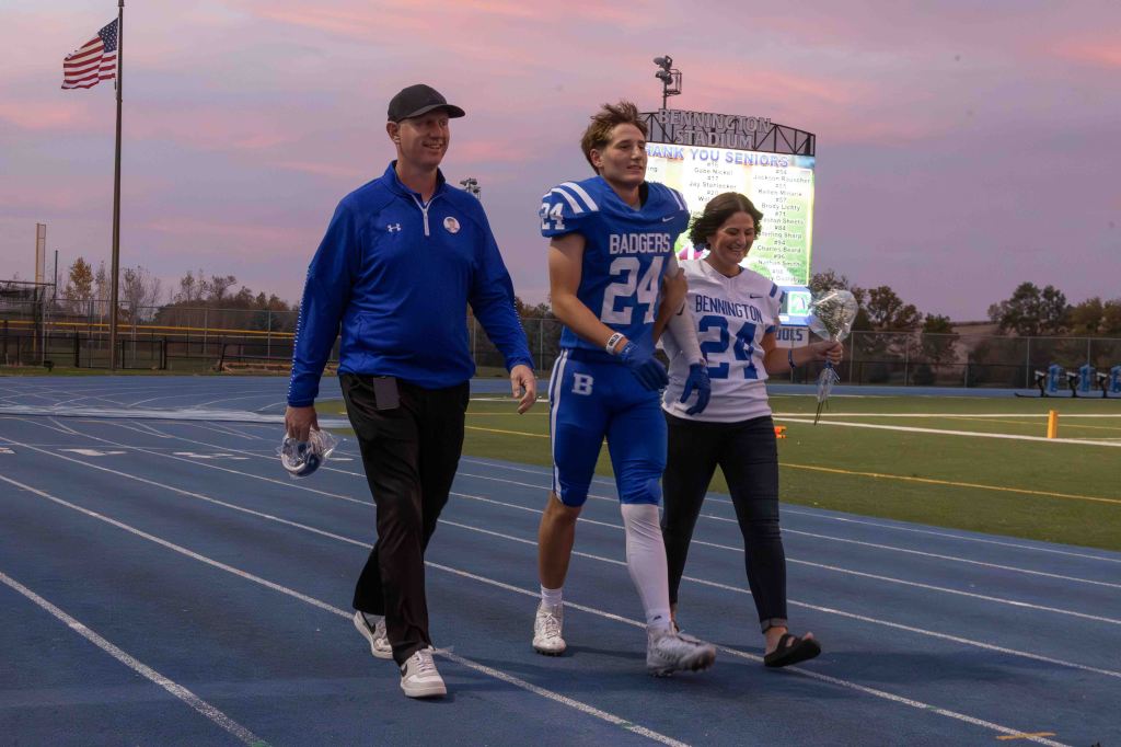A football player in blue and white uniform walking with a coach and a woman, holding flowers, on a track field during sunset.