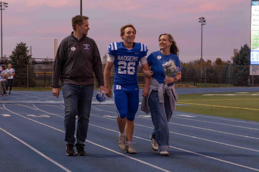 A high school football player in a blue uniform, number 26, smiling and walking hand-in-hand with his parents on a track during a sunset. One parent holds a bouquet of flowers.