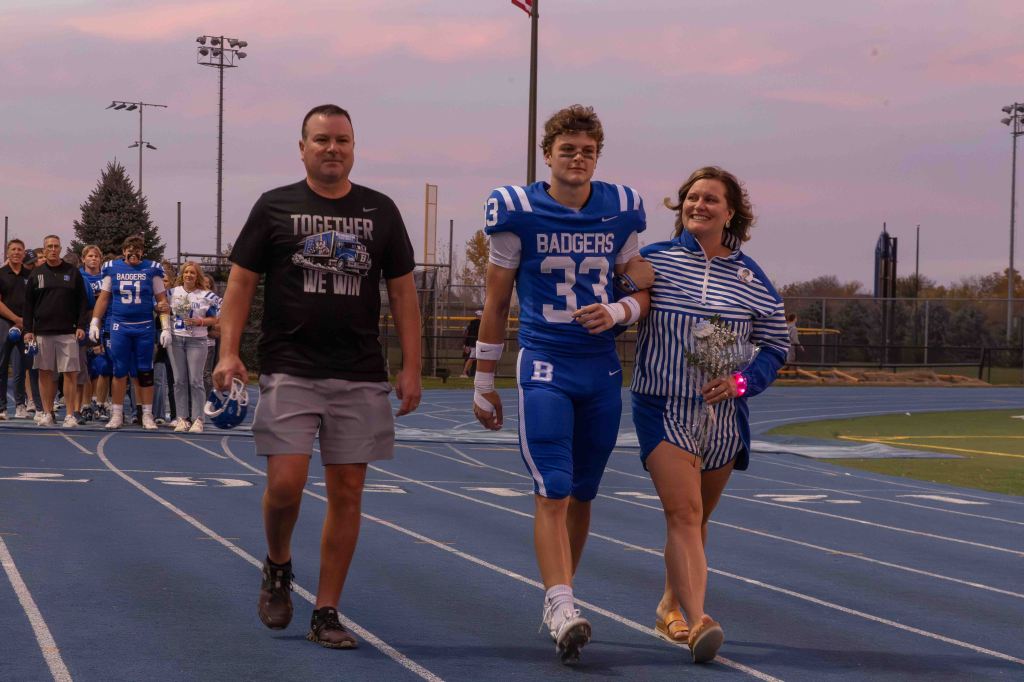 A football player wearing a blue jersey with the number 33 walks on the track with a woman holding flowers, accompanied by a man in a black shirt. A group of football players can be seen in the background.