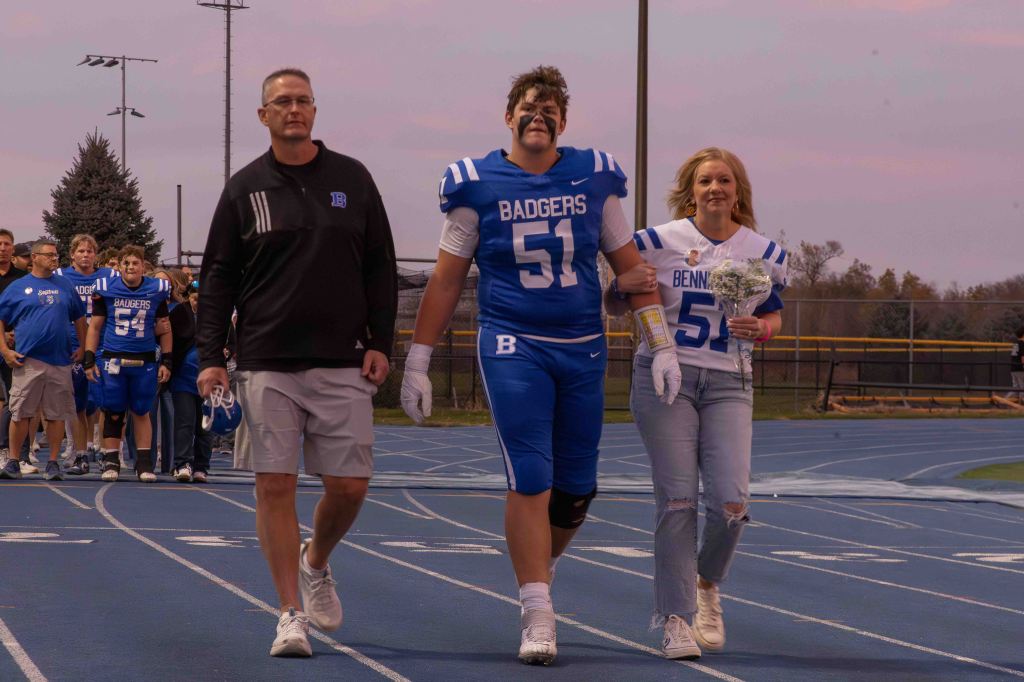 A senior football player in a blue jersey with 'Badgers' written on it walks on a track holding hands with a woman in a white jersey who is carrying flowers. They are escorted by a coach and a group of teammates.