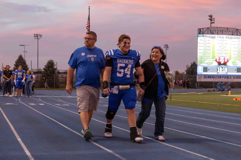 A senior football player in a blue uniform walks on the track with his parents during a recognition event. The sky is a mix of pink and purple hues at sunset.