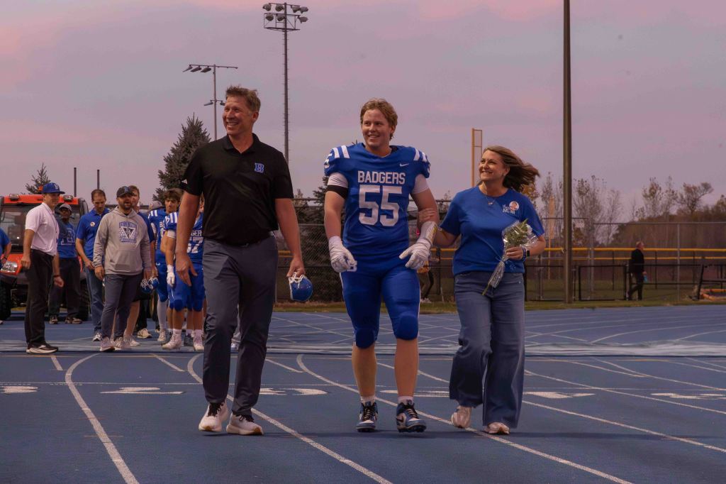 A football player in blue uniform number 55 walks on a track with two adults, a man in a black shirt and a woman in a blue shirt, during a sports event.