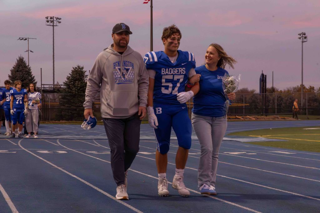 A high school football player in a blue jersey, number 57, is walking on a track surrounded by family. He is flanked by a woman holding flowers and a man wearing a Bennington Football hoodie. The sunset adds a warm glow to the scene, and other players can be seen in the background.
