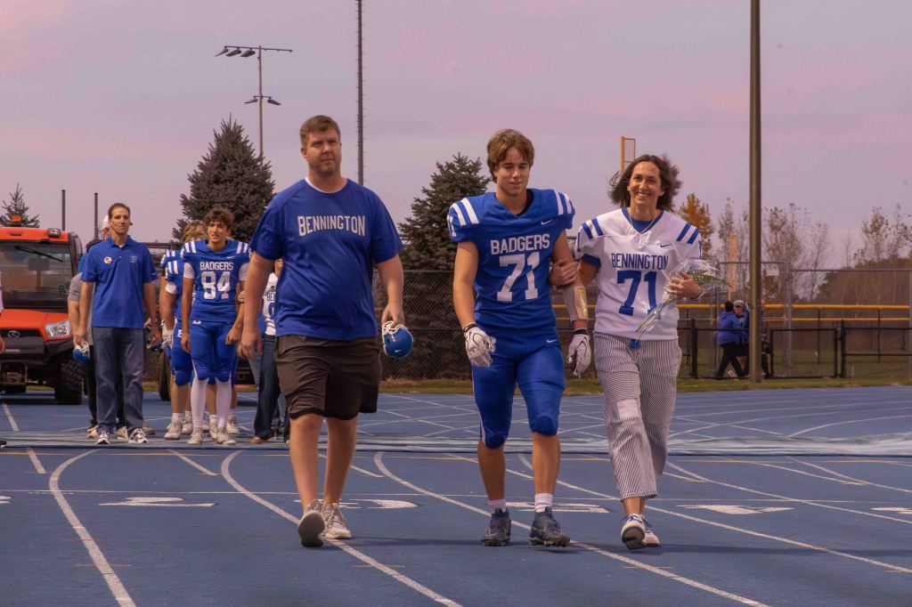 A football player wearing blue jersey number 71 walks on a blue track, accompanied by a woman in a white jersey and a man in a blue shirt. They are part of a sports event, with other team members visible in the background.