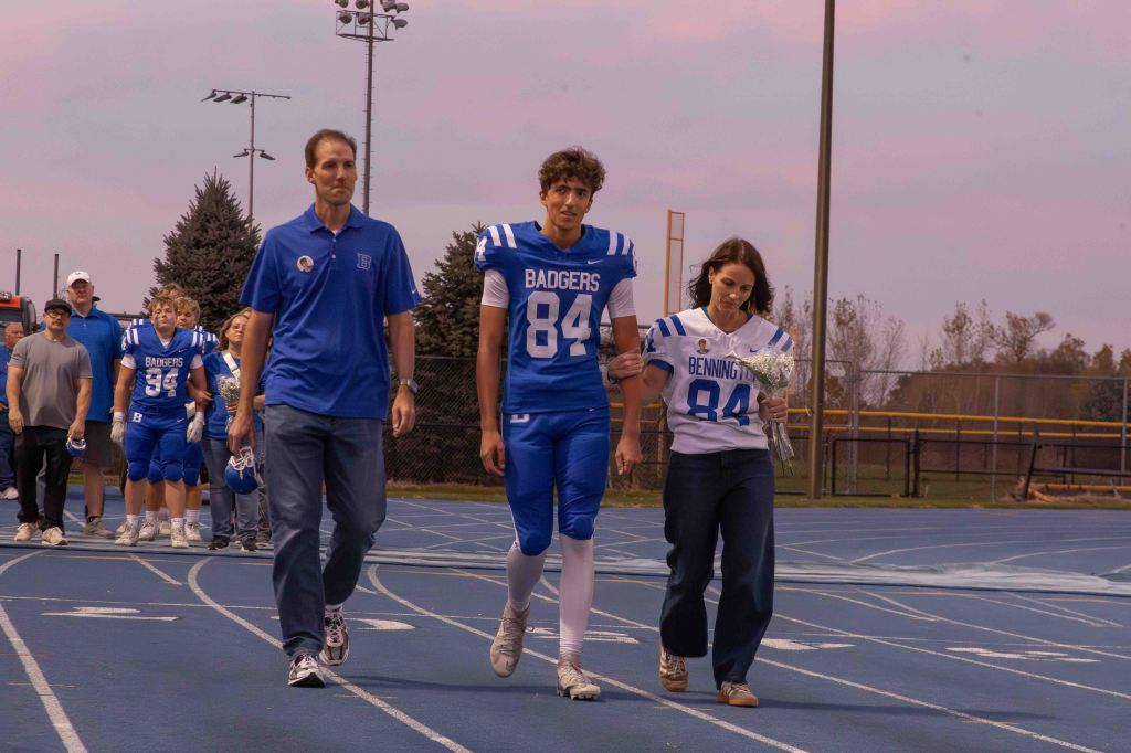 A high school football player in a blue uniform, flanked by his parents, walks on the track during a recognition event. The mother holds a bouquet of flowers, and the group is surrounded by teammates in the background.