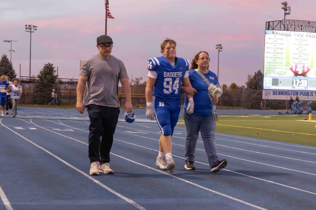 A young football player in a blue jersey walks on a track with his parents, one holding a bouquet of flowers, during a sports event at a stadium.