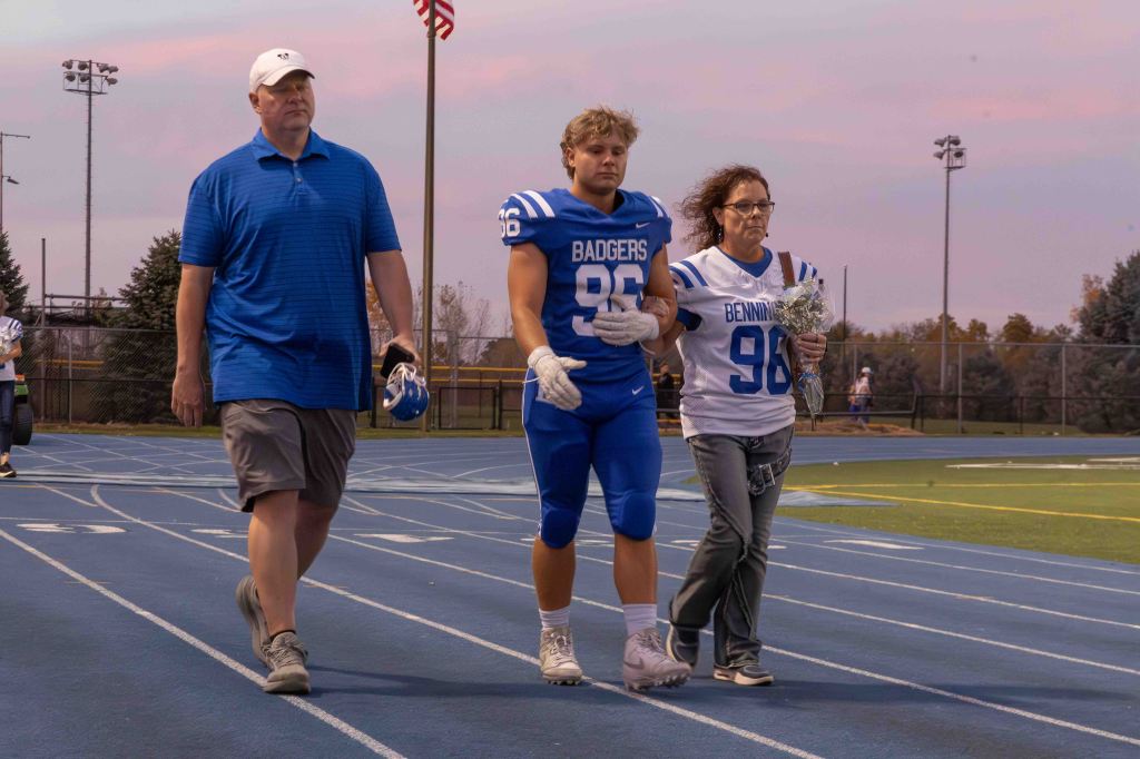 A high school football player wearing a blue jersey walks on the field with his parents, one wearing a blue polo shirt and the other in a white jersey, holding flowers.