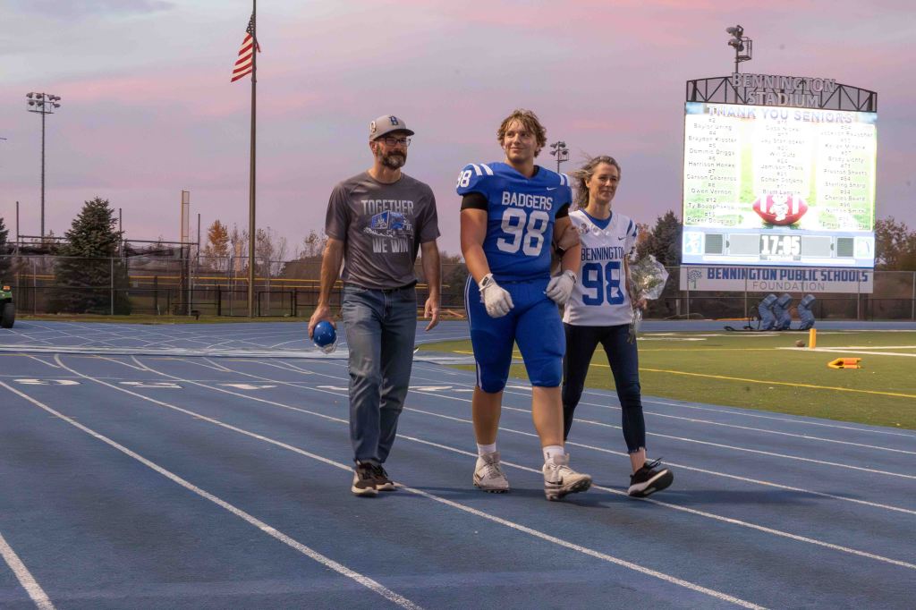 A football player in a blue jersey walks on a track, flanked by a man wearing a gray shirt and a woman in a white jersey, both holding a blue football. The background features a scoreboard and an American flag.