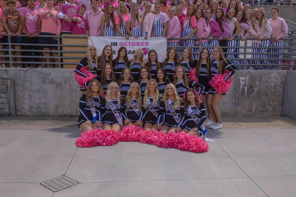 A group of cheerleaders in black and blue uniforms with pink pom-poms posing in front of a crowd, some wearing pink attire, holding a sign that reads 'IN OCTOBER WE WEAR PINK'.
