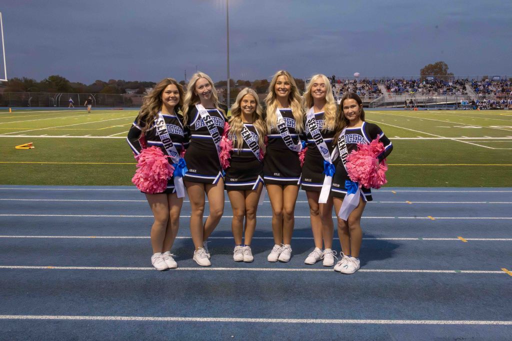 A group of six cheerleaders posing together on a football field, wearing cheerleading uniforms and holding pink pom-poms.
