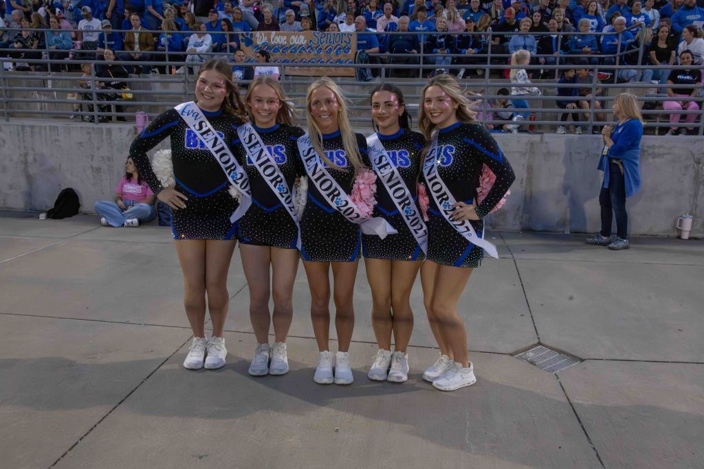 Five cheerleaders posing together in matching outfits and 'Senior 2023' sashes at a school event, with a crowd in the background.