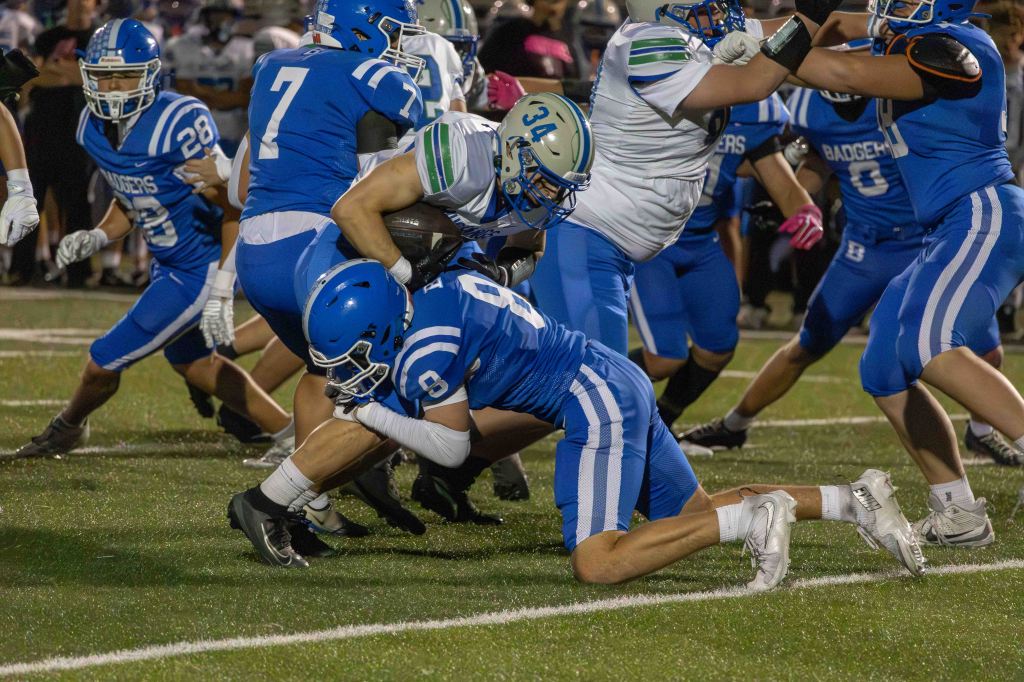 A high school football game with several players in blue and white uniforms attempting to tackle an opponent carrying the ball. The scene depicts action on the field with players engaged in a competitive play.