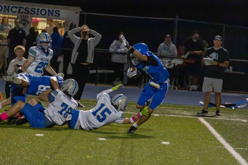 A football player in a blue uniform evades a tackle as two opposing players in white and blue uniforms attempt to bring him down during a night game.