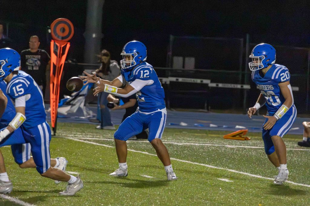 A youth football player in a blue uniform, wearing jersey number 12, is preparing to pass the ball during a nighttime game, while teammates in the background are in motion.