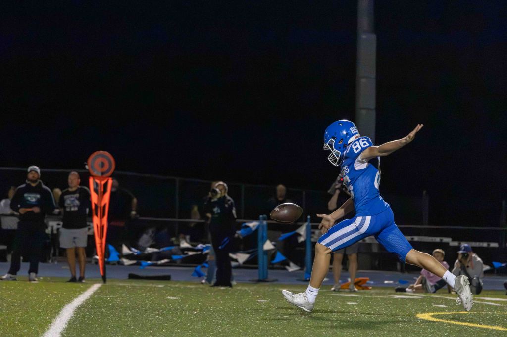 A football player in a blue uniform and helmet kicking a ball on a field, with spectators in the background during a night game.