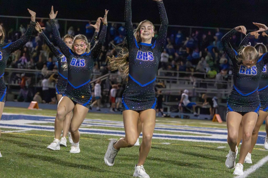 A group of cheerleaders in sparkling uniforms performing at a nighttime sports event, with the crowd in the background.